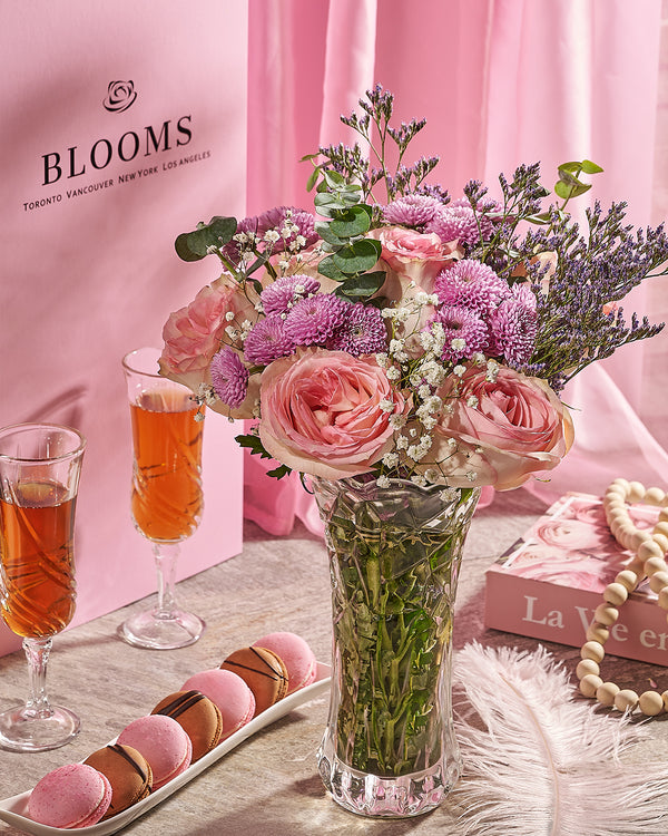 Bouquet of pink and purple flowers in a vase with two glasses of tea and pastries on a table against a pink background.