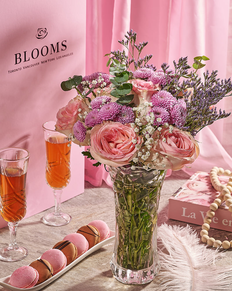 Bouquet of pink and purple flowers in a vase with two glasses of tea and pastries on a table against a pink background.