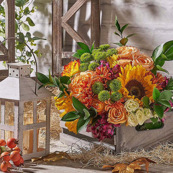 Colorful flower arrangement in a wooden crate with a lantern and pomegranates on a rustic wooden surface.