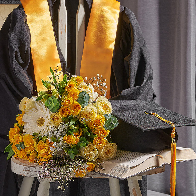 Graduation gown with yellow stole and floral arrangement on a stool.