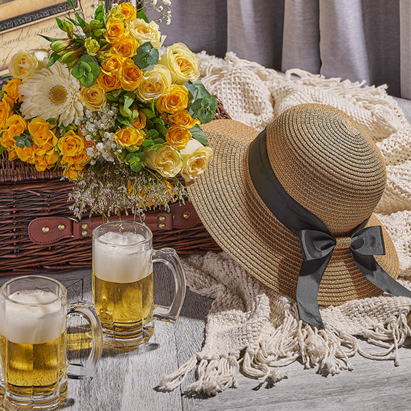 Straw hat with a black ribbon, two beer mugs, and a basket of flowers on a wooden table.