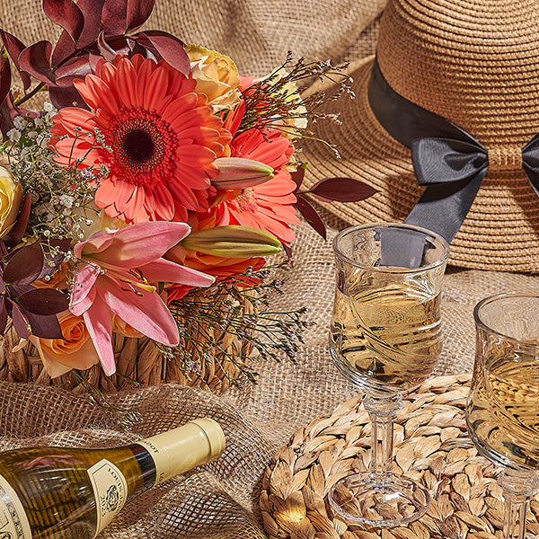 Wine bottle, glasses, and floral arrangement on a textured surface with a straw hat.