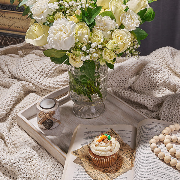 Bouquet of white and yellow flowers in a glass vase on a wooden tray with a cupcake and book underneath.