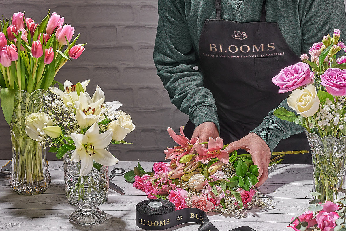 Person arranging flowers with 'BLOOMS' branding in an apron, on a table with floral arrangements.