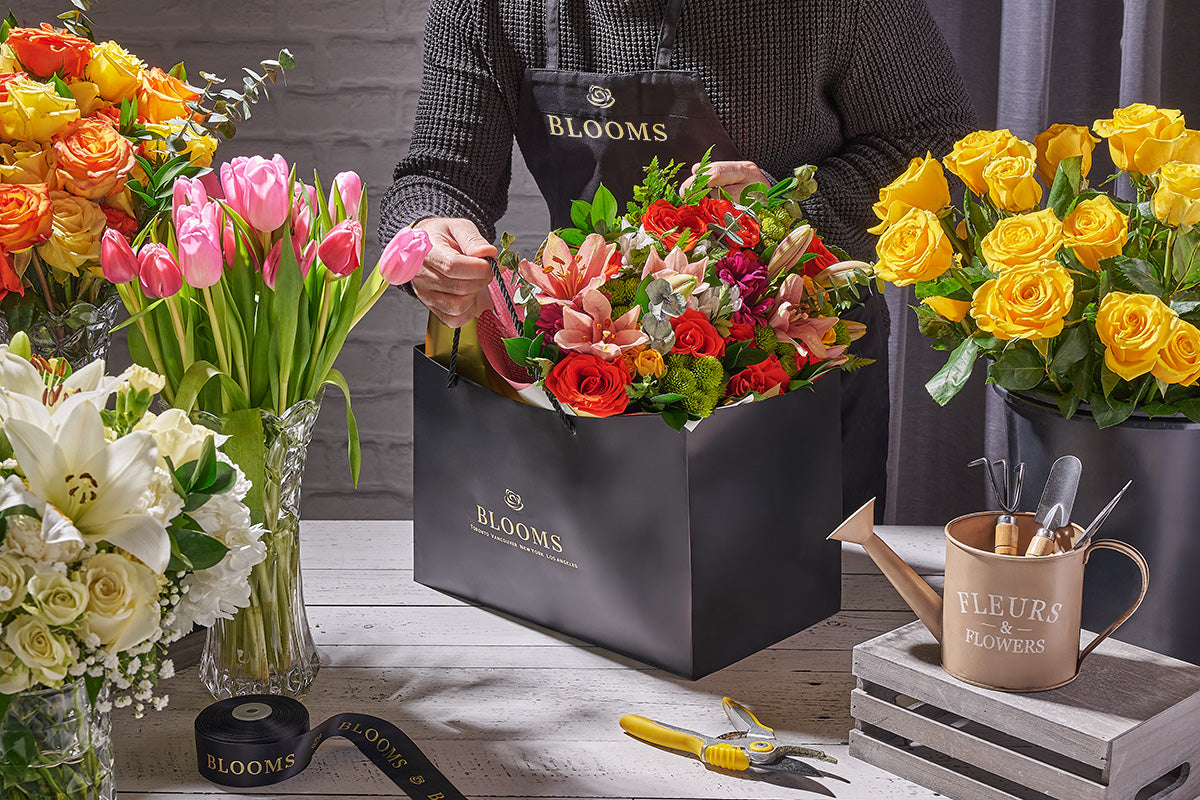 Florist packaging a bouquet of flowers in a 'BLOOMS' box with various floral arrangements and tools on a table.
